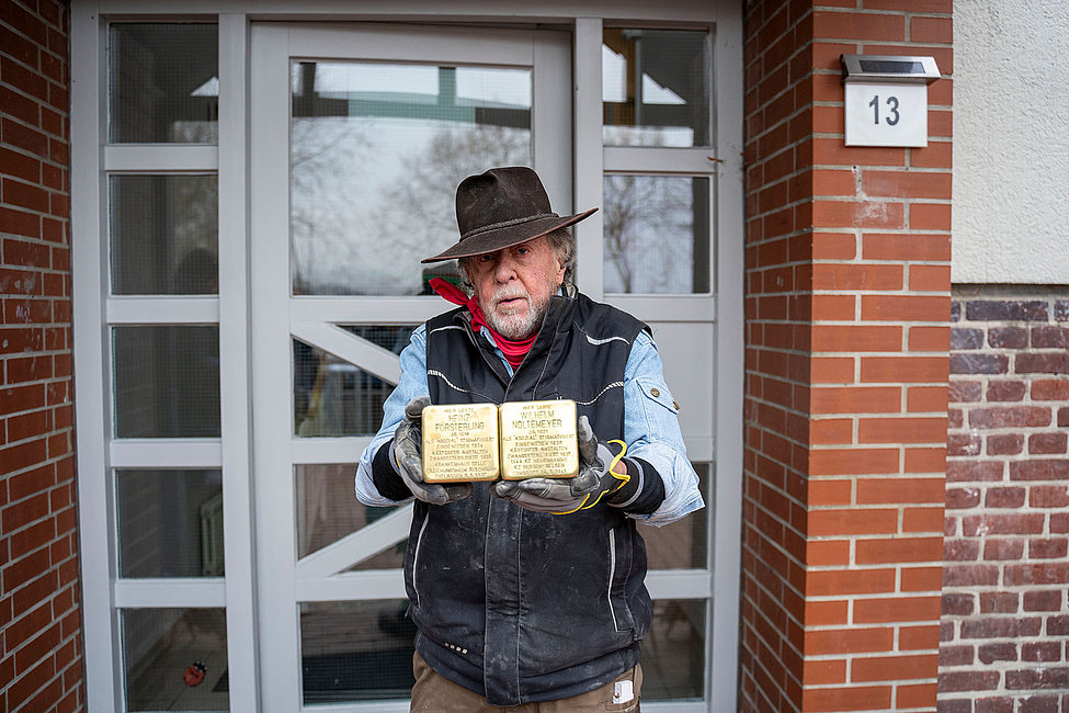 Mann mit Hut hält zwei Stolpersteine mit Inschriften vor Haustür